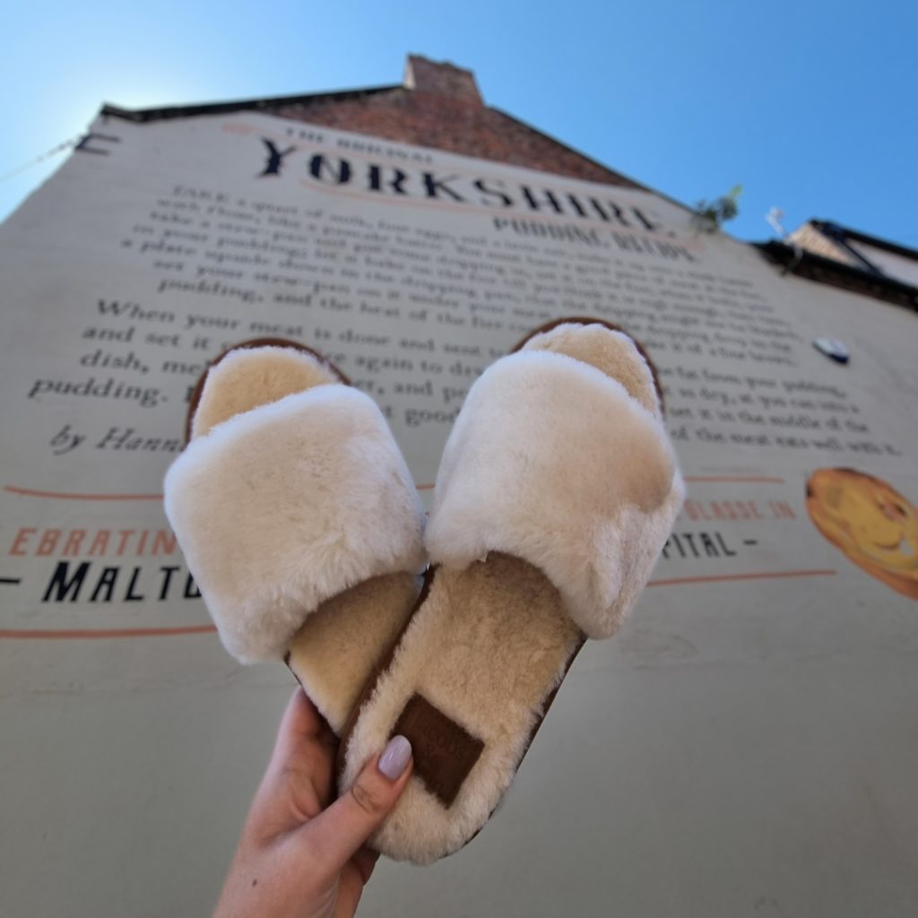 Fluffy cream slider style slippers held in the air in front of Malton's Yorkshire Pudding mural